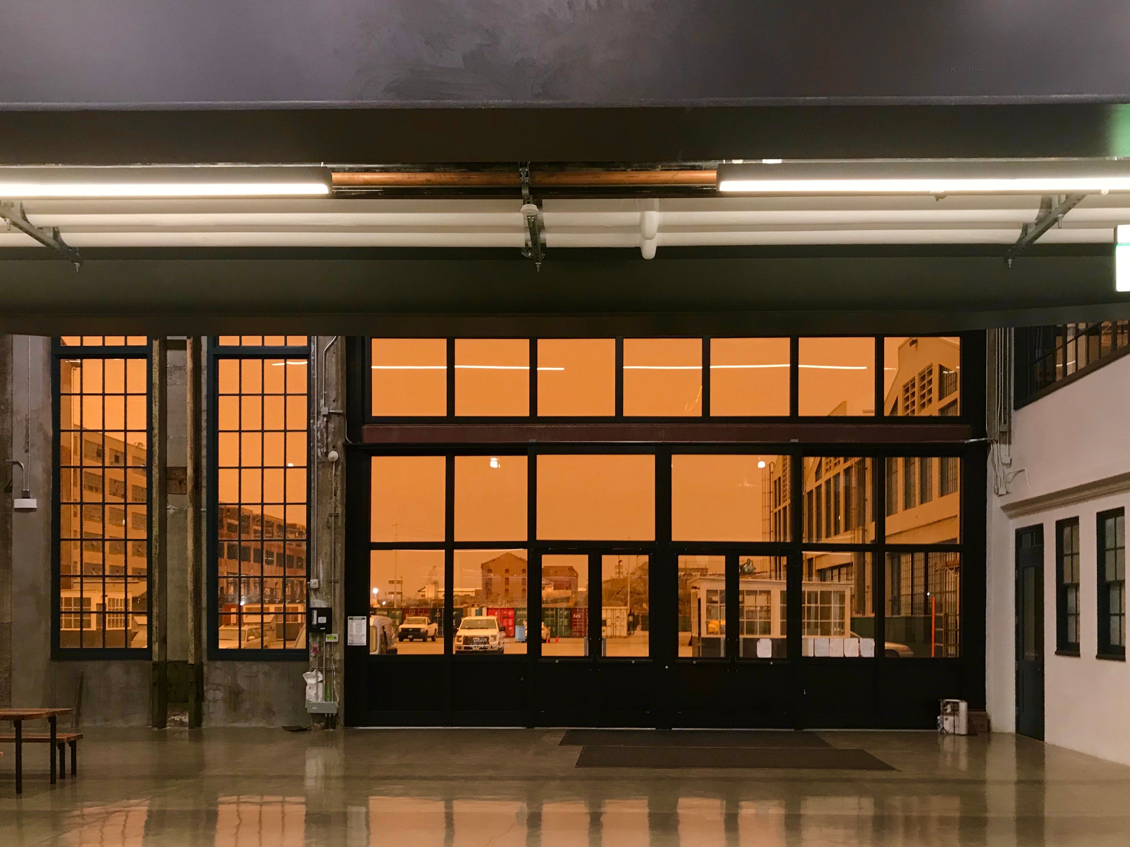 A photo inside a building looking out at a parking lot and sky. The sky is shockingly, saturatedly orange. The inside of the building is clear lit in a white bordering on austere.